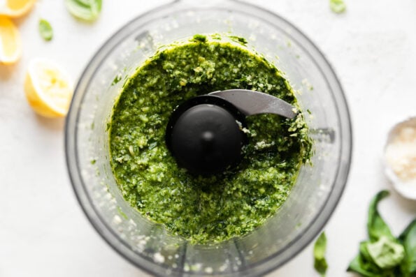 Blended lemon basil pesto in a food processor carafe, atop a white surface surrounded by additional pesto ingredients that are out of focus.