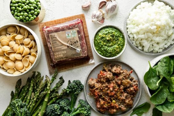 Lamb pasta ingredients arranged on a creamy white surface: spicy lamb sausage made with New Zealand grass-fed ground lamb, garlic, pasta, asparagus, broccolini, baby spinach, yellow onion, English peas, and lemon basil pesto.