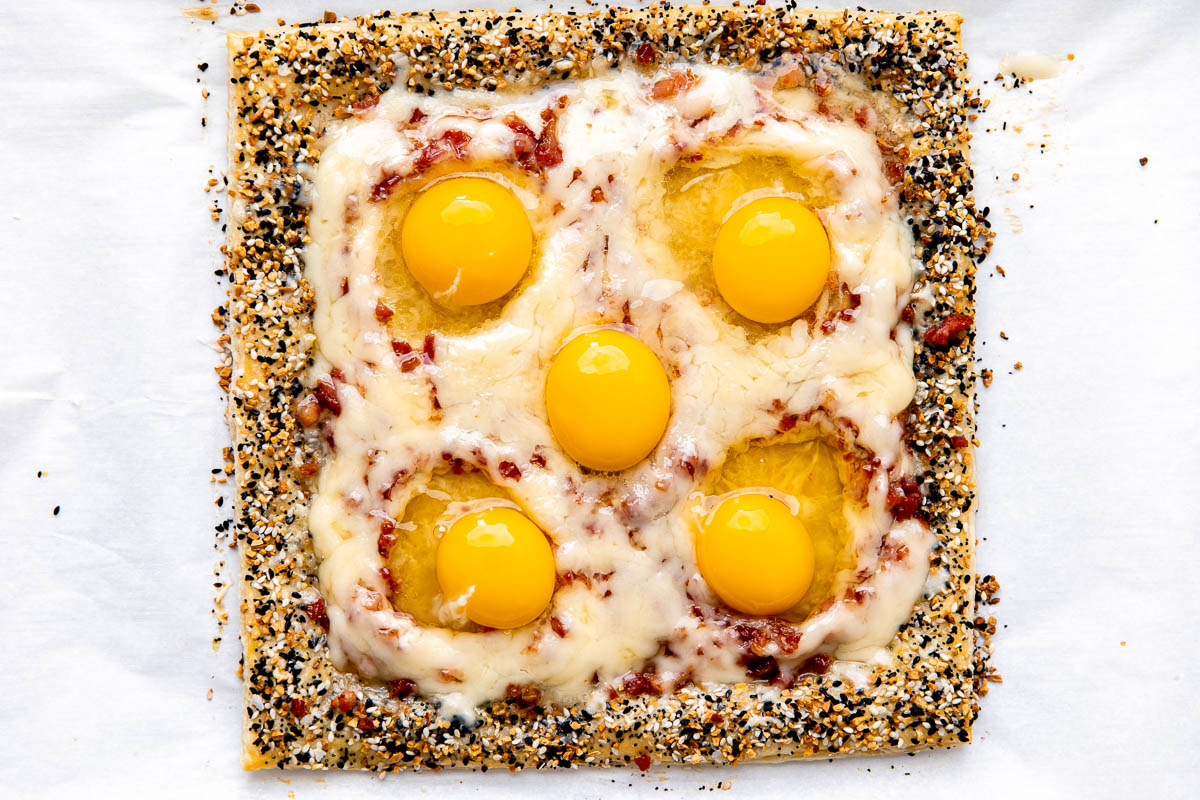Five uncooked eggs have been placed inside of prepared wells within a par-baked breakfast pizza. The pizza sits atop a parchment lined baking sheet.