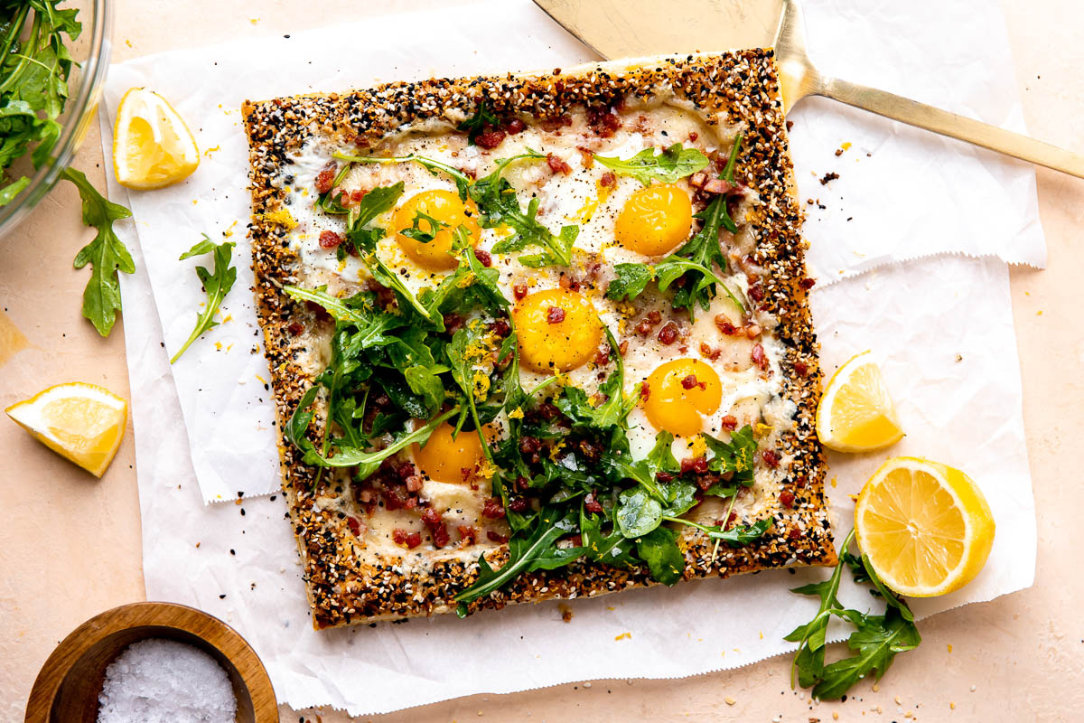 A finished puff pastry breakfast pizza is served atop a piece of parchment paper and rests atop a peach colored textured surface. The pizza has been garnished with arugula lightly dressed in olive oil, lemon zest, kosher salt, and ground black pepper. Quartered and halved pieces of lemon along with loose arugula leaves surround the pizza along with a small wooden pinch bowl filled with kosher salt, a clear glass bowl filled with arugula, and a gold serving utensil.
