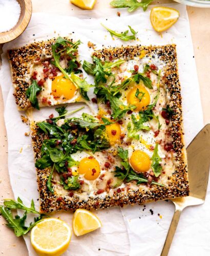 A finished puff pastry breakfast pizza is served atop a piece of parchment paper and rests atop a peach colored textured surface. The pizza has been garnished with arugula lightly dressed in olive oil, lemon zest, kosher salt, and ground black pepper. Quartered and halved pieces of lemon along with loose arugula leaves surround the pizza along with a small wooden pinch bowl filled with kosher salt, a clear glass bowl filled with arugula, and a gold serving utensil. A slice of pizza has been cut for serving and is slightly separated from the rest of the breakfast pizza.