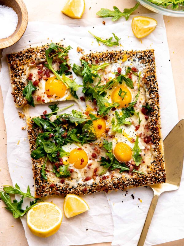 A finished puff pastry breakfast pizza is served atop a piece of parchment paper and rests atop a peach colored textured surface. The pizza has been garnished with arugula lightly dressed in olive oil, lemon zest, kosher salt, and ground black pepper. Quartered and halved pieces of lemon along with loose arugula leaves surround the pizza along with a small wooden pinch bowl filled with kosher salt, a clear glass bowl filled with arugula, and a gold serving utensil. A slice of pizza has been cut for serving and is slightly separated from the rest of the breakfast pizza.