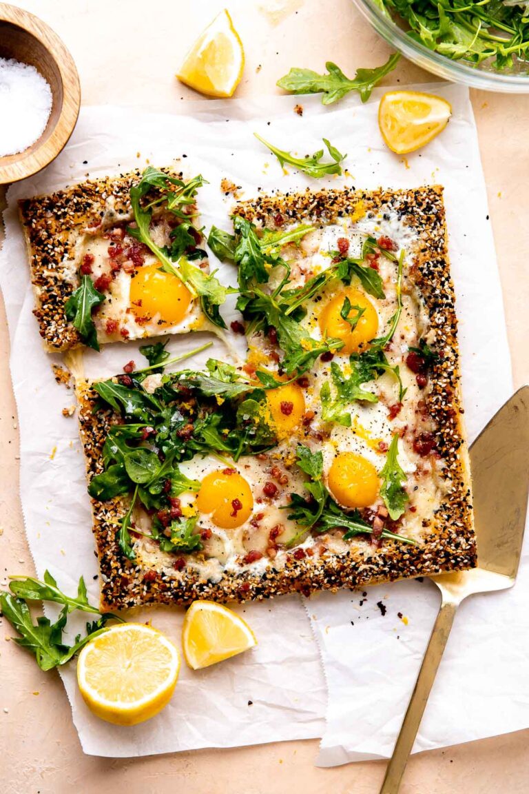 A finished puff pastry breakfast pizza is served atop a piece of parchment paper and rests atop a peach colored textured surface. The pizza has been garnished with arugula lightly dressed in olive oil, lemon zest, kosher salt, and ground black pepper. Quartered and halved pieces of lemon along with loose arugula leaves surround the pizza along with a small wooden pinch bowl filled with kosher salt, a clear glass bowl filled with arugula, and a gold serving utensil. A slice of pizza has been cut for serving and is slightly separated from the rest of the breakfast pizza.