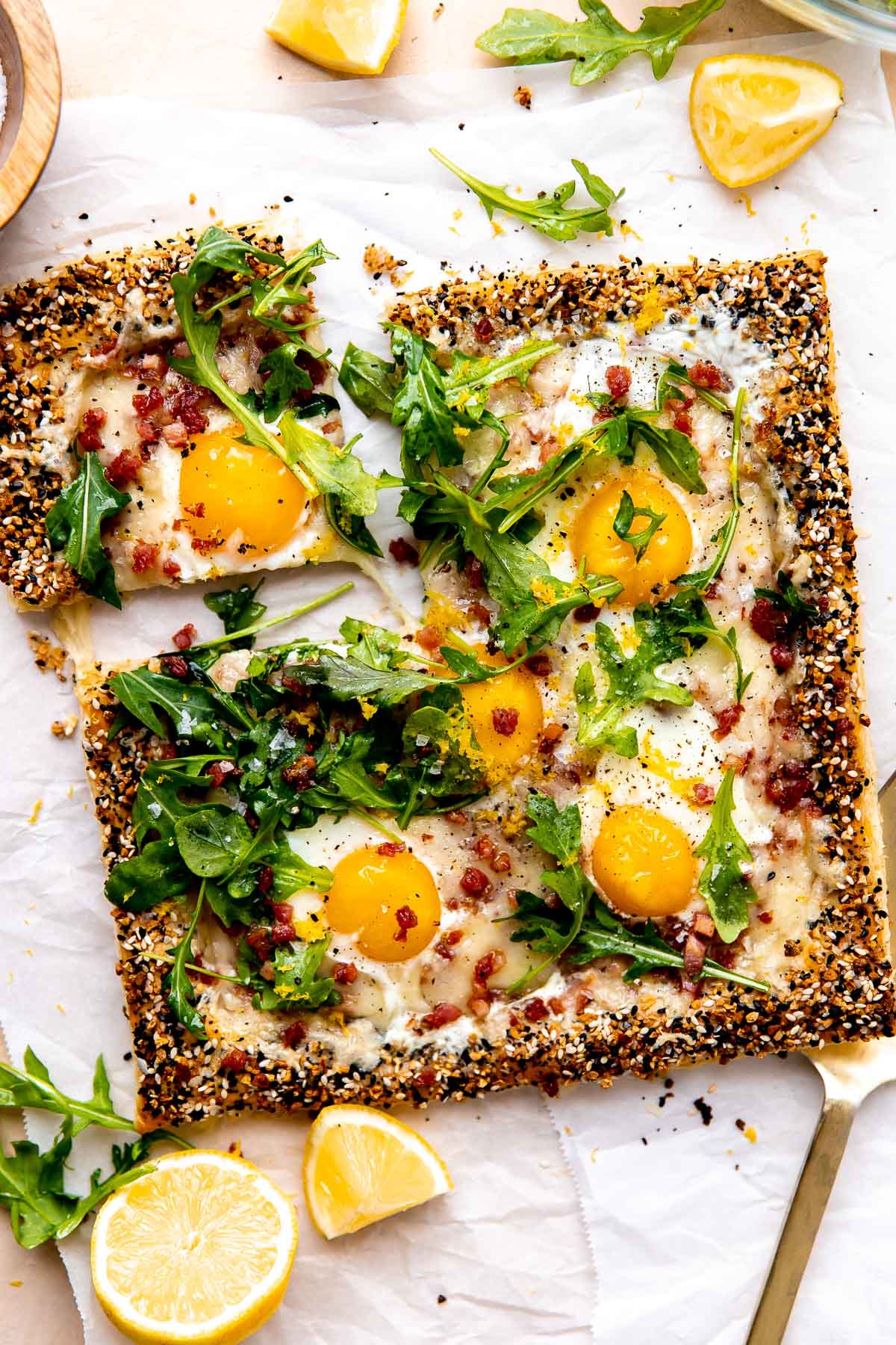 A finished breakfast pizza is served atop a piece of parchment paper and rests atop a peach colored textured surface. The pizza has been garnished with arugula lightly dressed in olive oil, lemon zest, kosher salt, and ground black pepper. Quartered and halved pieces of lemon along with loose arugula leaves surround the pizza along with a small wooden pinch bowl filled with kosher salt, a clear glass bowl filled with arugula, and a gold serving utensil. A slice of pizza has been cut for serving and is slightly separated from the rest of the breakfast pizza.