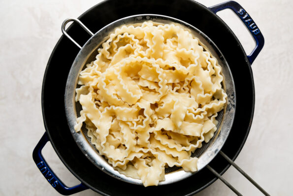 How to make mushroom ragu pasta, step 4: boil the pasta. A fine mesh strainer filled with drained mafaldine pasta rests atop a dark blue Staub cocotte. The cocotte sits atop a creamy white textured surface.