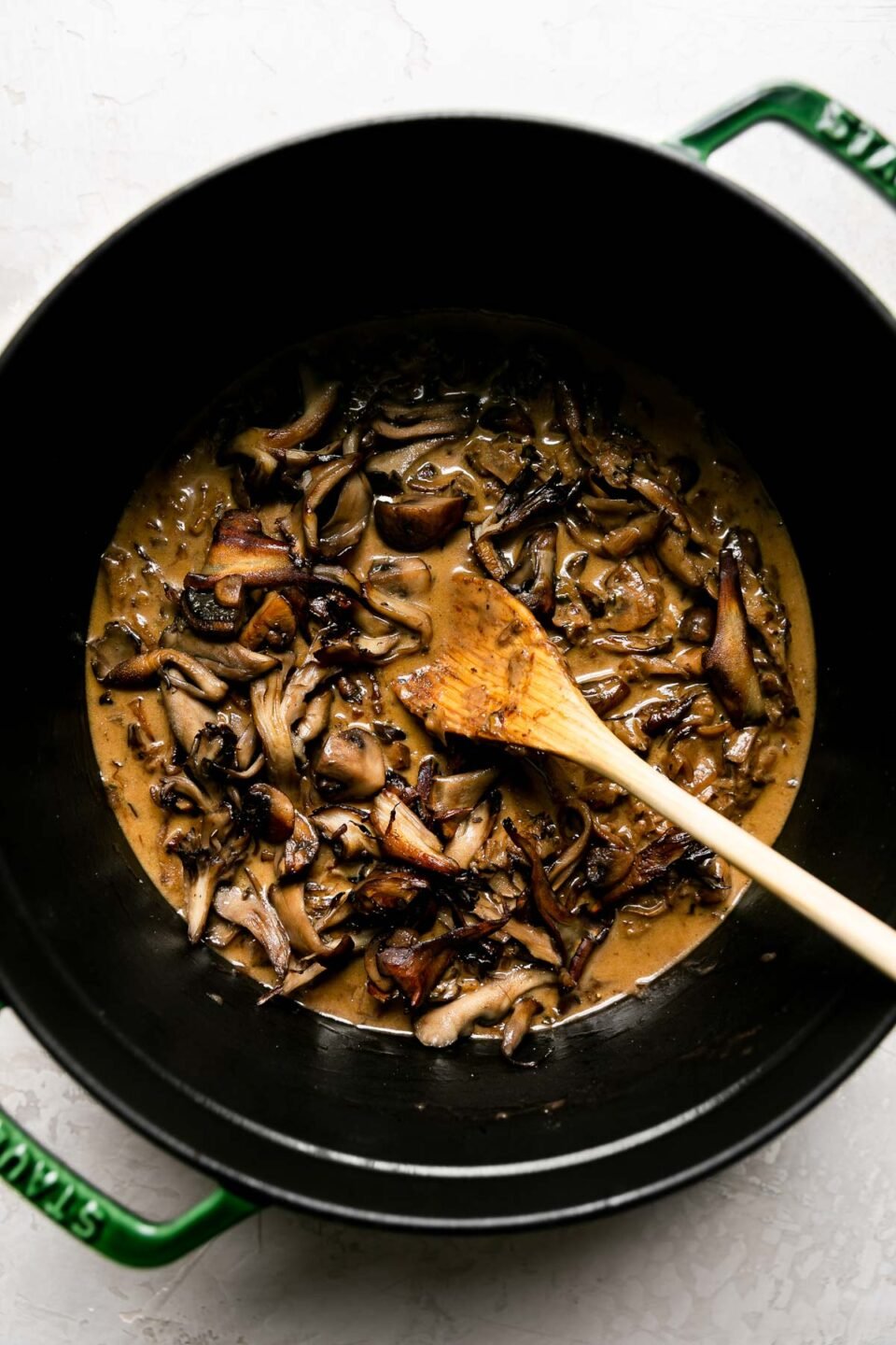 How to make mushroom ragu sauce, step 6: build & simmer the mushroom ragu. A porcini-infused stock rests inside of a green Staub cocotte. Heavy cream & browned fresh mushrooms are added to the pot & a wooden spoon rests inside of the pot to help build the mushroom ragu. The cocotte sits atop a creamy textured surface.