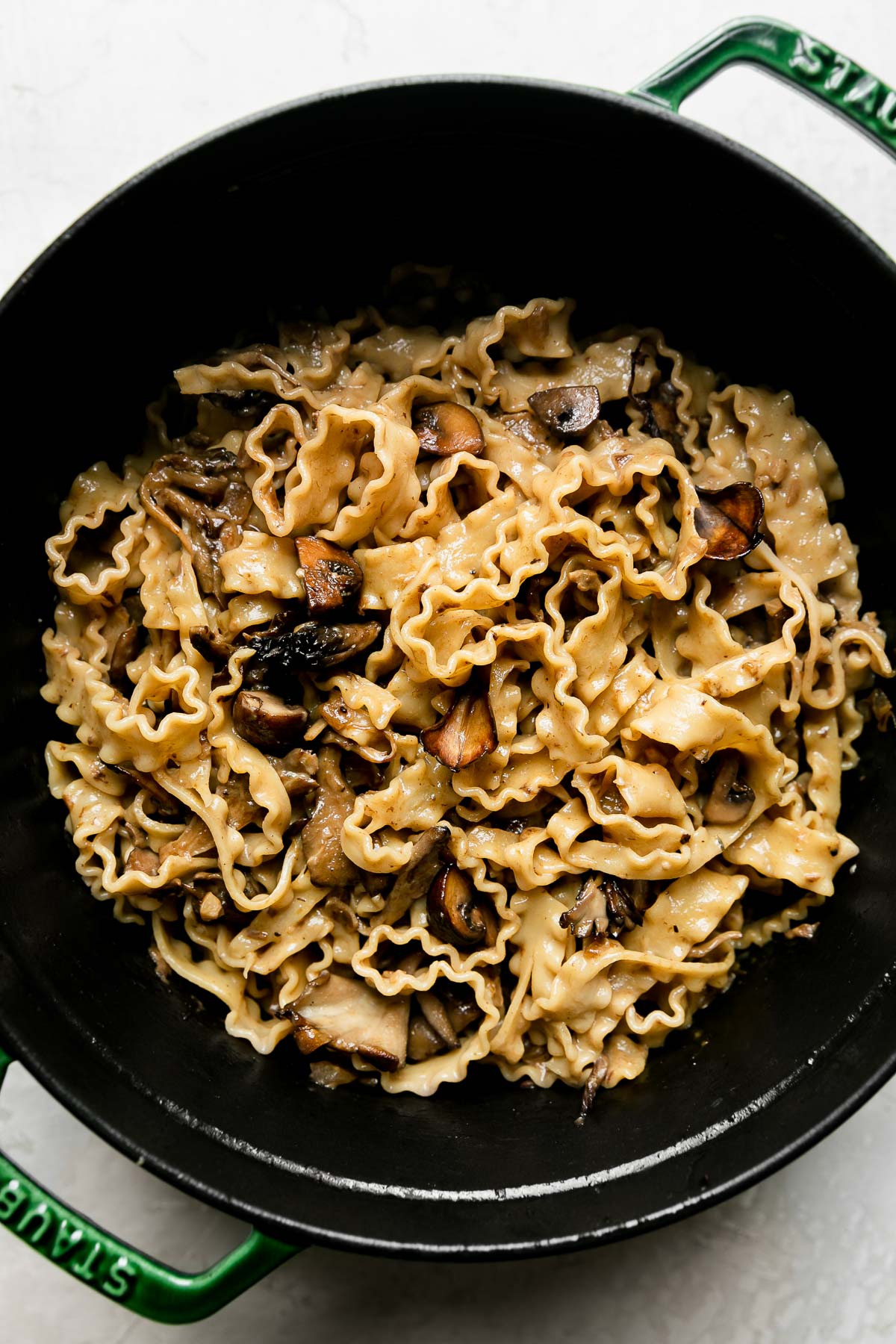 How to make wild mushroom ragu, step 7: finish the mushroom ragu pasta. Cooked pasta is added to & tossed with a wild mushroom ragu sauce in a green Staub cocotte. The cocotte sits atop a creamy white textured surface.