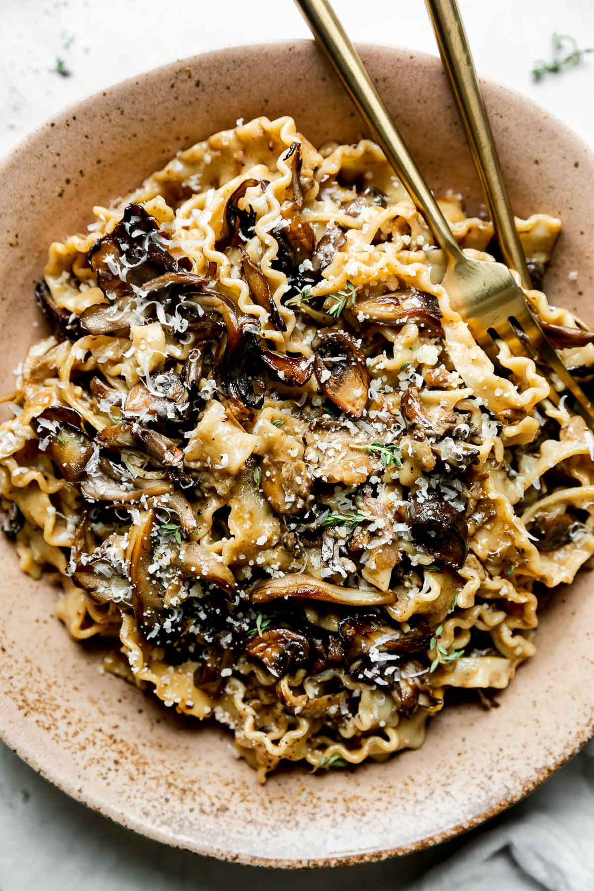 A serving of wild mushroom ragu pasta fills a brown speckled ceramic bowl. The pasta has been garnished with freshly grated parmesan and herbs. The bowl sits atop a creamy white textured surface with a light gray linen napkin resting alongside and few fresh herbs sprinkled loosely nearby. Gold silverware rests inside of the bowl.