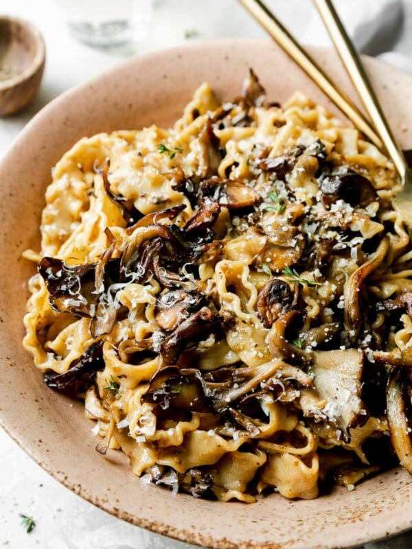A side angle shot of a serving of wild mushroom ragu pasta inside of a brown speckled ceramic bowl. The pasta has been garnished with freshly grated parmesan and herbs and has gold silverware resting inside of the bowl. The bowl sits atop a creamy white textured surface with a light gray linen napkin, a small wooden pinch bowl filled with black pepper, and a glass of white wine resting alongside.