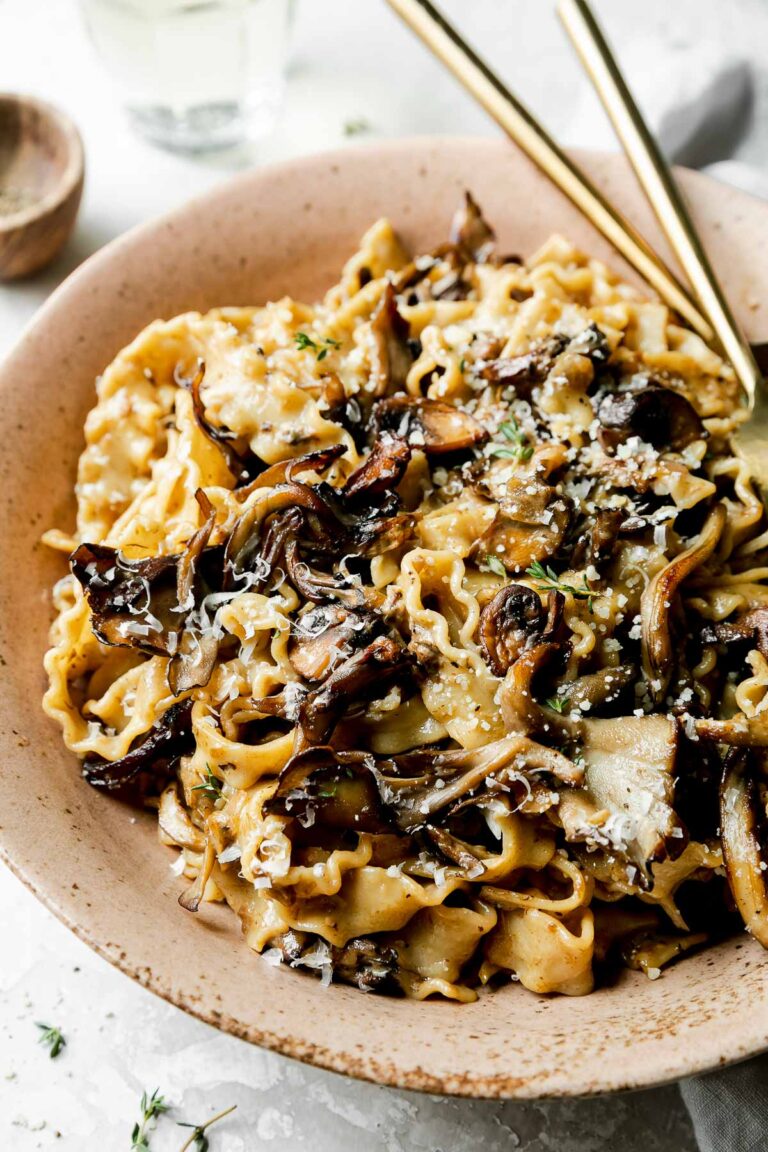 A side angle shot of a serving of wild mushroom ragu pasta inside of a brown speckled ceramic bowl. The pasta has been garnished with freshly grated parmesan and herbs and has gold silverware resting inside of the bowl. The bowl sits atop a creamy white textured surface with a light gray linen napkin, a small wooden pinch bowl filled with black pepper, and a glass of white wine resting alongside.