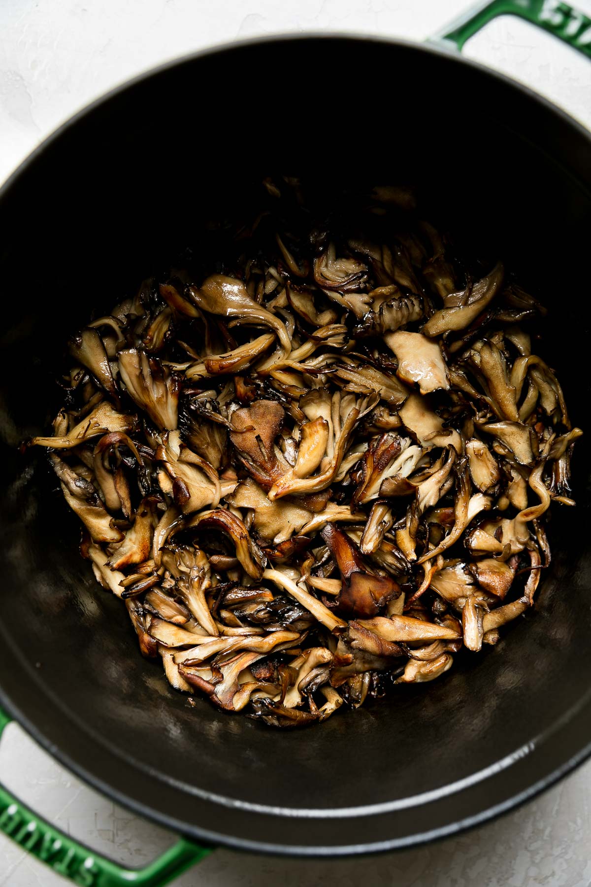 How to make wild mushroom ragu step 1: fresh mushrooms brown in the bottom of a green Staub cocotte. The cocotte sits atop a creamy white textured surface.