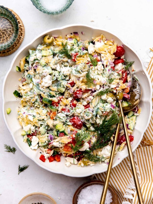 A large white bowl with scalloped edges is filled assembled and mixed greek orzo pasta salad garnished with fresh dill. The bowl sits atop a creamy white textured surface with a yellow and white linen napkin partially tucked underneath on one side. Two blue glasses rest alongside the bowl at center, one rests atop a wicker coaster. While a small ceramic pinch bowl filled with ground black pepper and a small wooden pinch bowl filled with kosher salt rest below the bowl at center. Two gold serving spoons rest inside of the greek pasta salad.