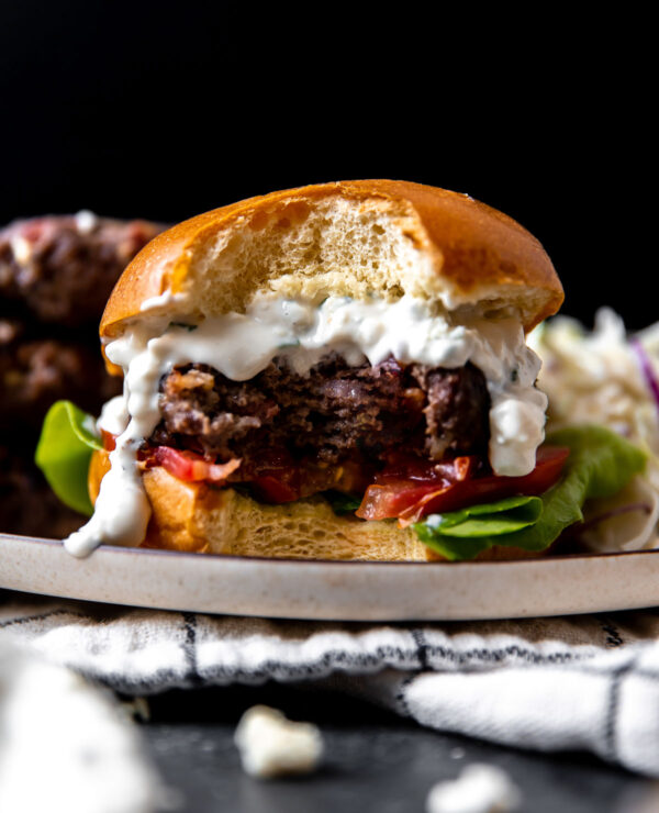 A side angle shot of a finished grilled blue cheese burger atop a plate with a large bite taken out of it. The plate sits atop a white and black window-pane patterned linen napkin that sits atop a dark gray textured surface. The burger is sandwiched between a burger bun with sliced tomato and lettuce sandwiched underneath and with creamy blue cheese sauce spooned over top the burger patty. Three burger patties and crunch apple slaw sit alongside the burger on the same plate.