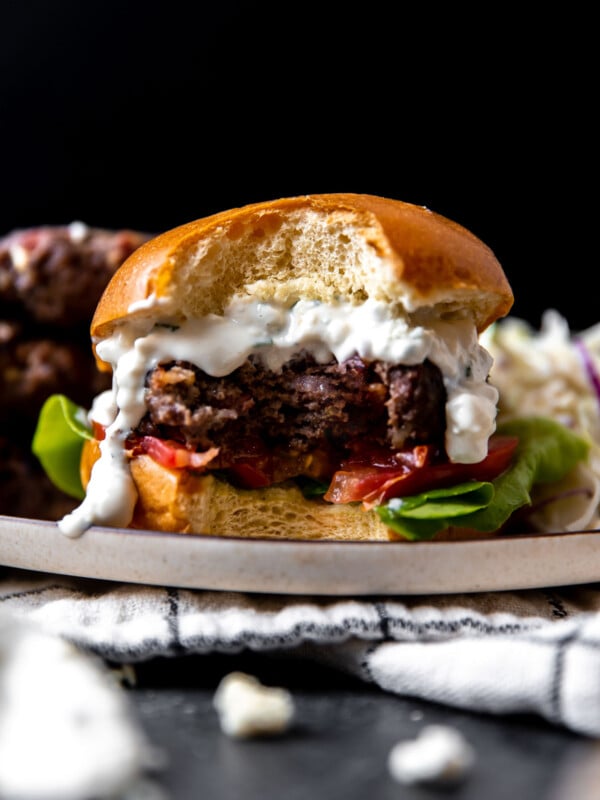 A side angle shot of a finished grilled blue cheese burger atop a plate with a large bite taken out of it. The plate sits atop a white and black window-pane patterned linen napkin that sits atop a dark gray textured surface. The burger is sandwiched between a burger bun with sliced tomato and lettuce sandwiched underneath and with creamy blue cheese sauce spooned over top the burger patty. Three burger patties and crunch apple slaw sit alongside the burger on the same plate.