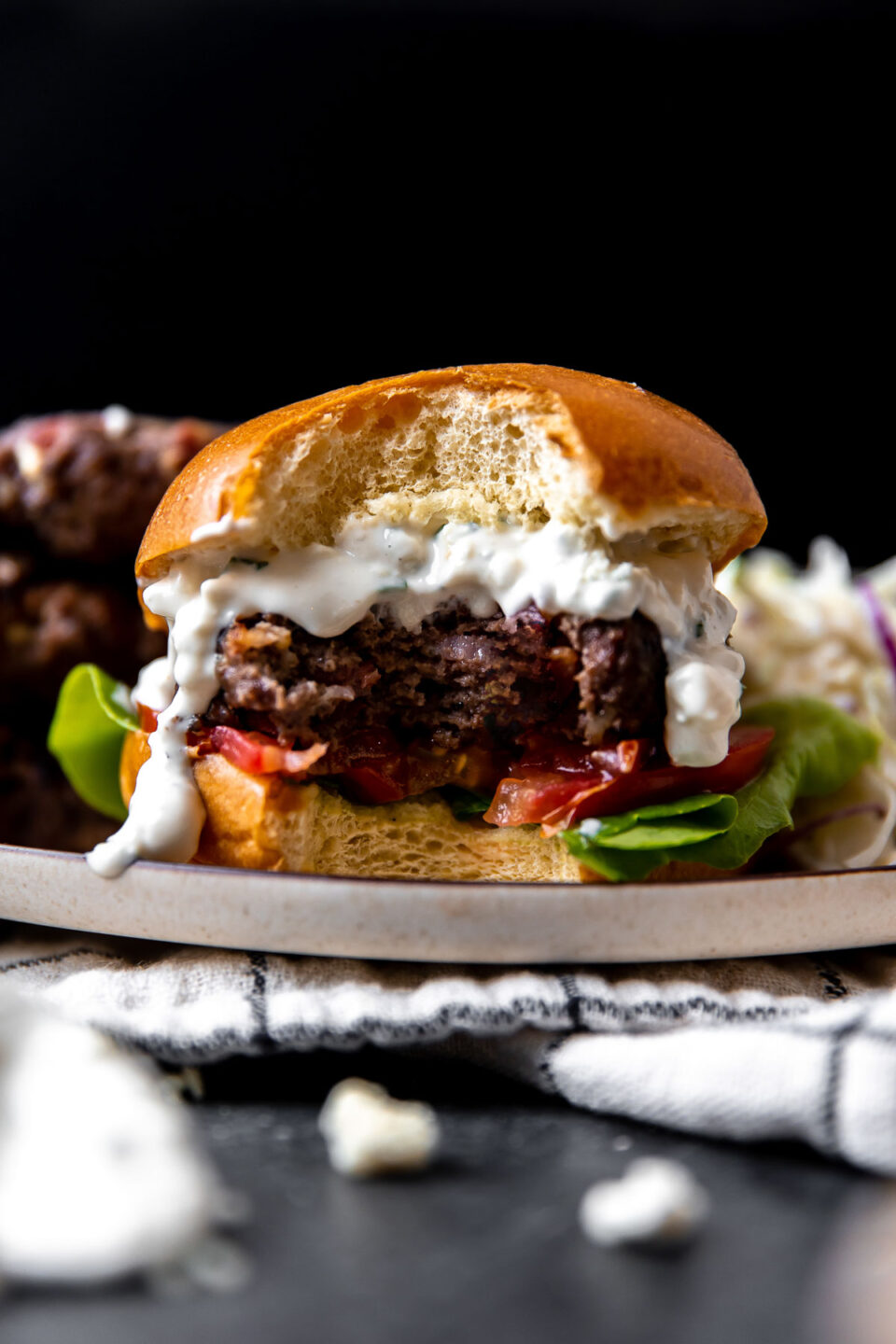A side angle shot of a finished grilled blue cheese burger atop a plate with a large bite taken out of it. The plate sits atop a white and black window-pane patterned linen napkin that sits atop a dark gray textured surface. The burger is sandwiched between a burger bun with sliced tomato and lettuce sandwiched underneath and with creamy blue cheese sauce spooned over top the burger patty. Three burger patties and crunch apple slaw sit alongside the burger on the same plate.