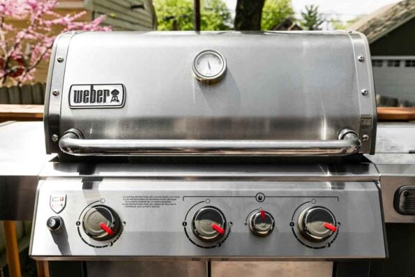 A Weber Genesis II stainless steel grill sits atop an outdoor deck. The grill preheats to prepare grilled hot dogs.