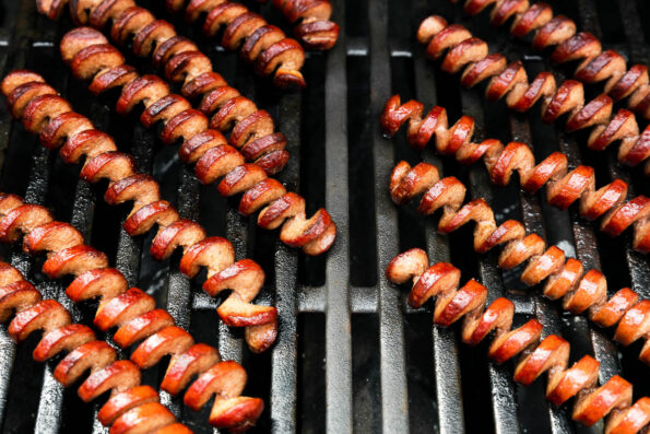 A slightly overhead shot of spiralized hot dogs being grilled atop gas grill grates.