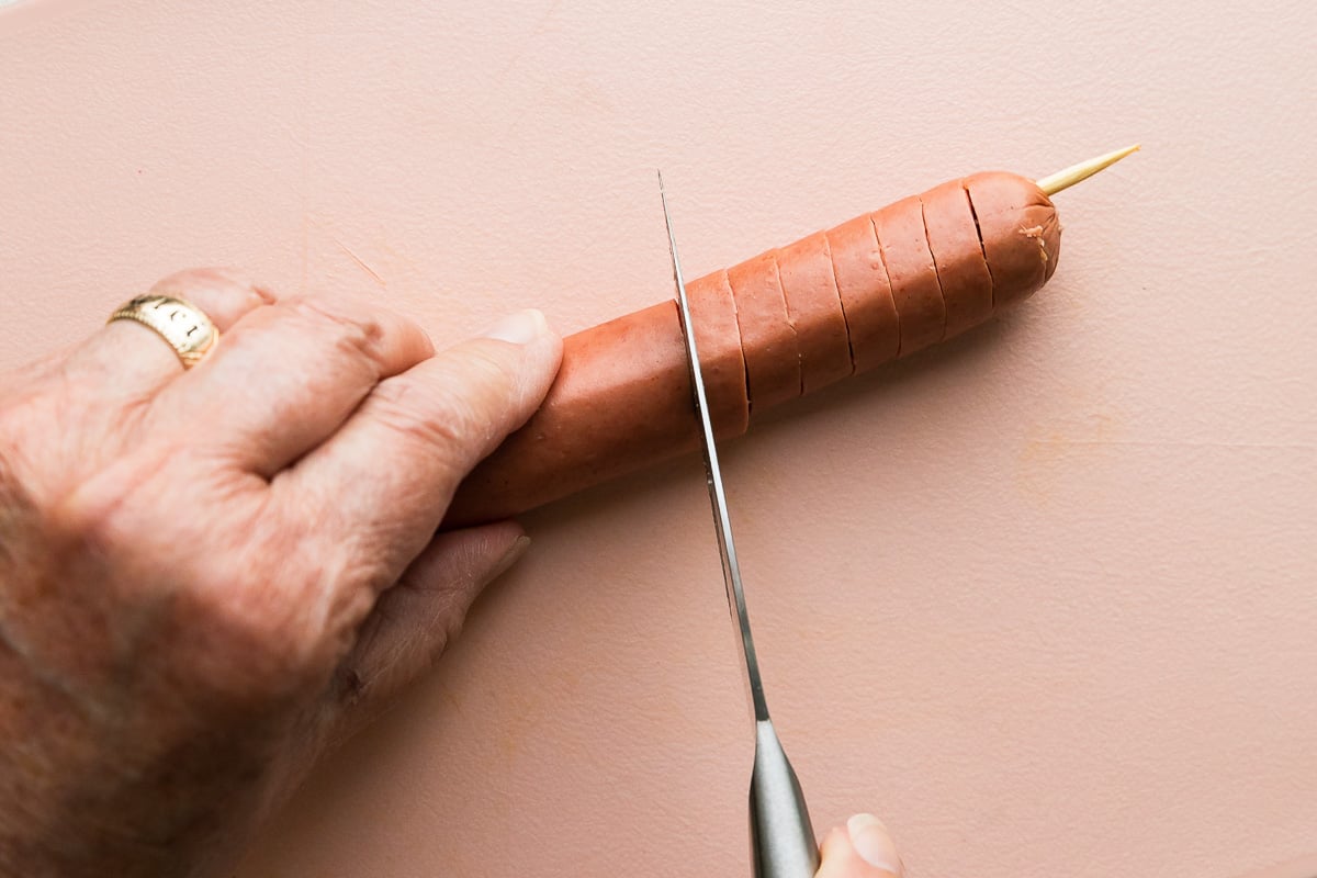 A woman stabilizes a single hot dog with a wooden skewer placed through it lengthwise on a plastic pink cutting board with one hand while holding a pairing knife in the other. She presses the blade of the knife through the hot dog until it hits the skewer, while rotating the skewer, to create a spiral cut hot dog. The cutting board sits atop a creamy white textured surface and a baking sheet full of spiral hot dogs rests just above the cutting board.