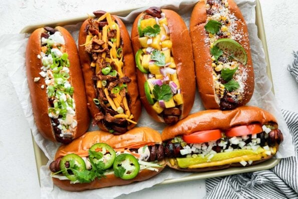 A variety of specialty grilled hot dogs are arranged atop a crumbled piece of parchment paper that lines an aluminum baking sheet, including: a Banh Mi hot dog, a Chicago-Style grilled hot dog, a Teriyaki-Style spiral hot dog, a BBQ-Style hot dog, a Buffalo-Style grilled hot dog, and an Elote spiral hot dog. The baking sheet sits atop a creamy white textured surface and a blue and white striped linen napkin rests alongside.