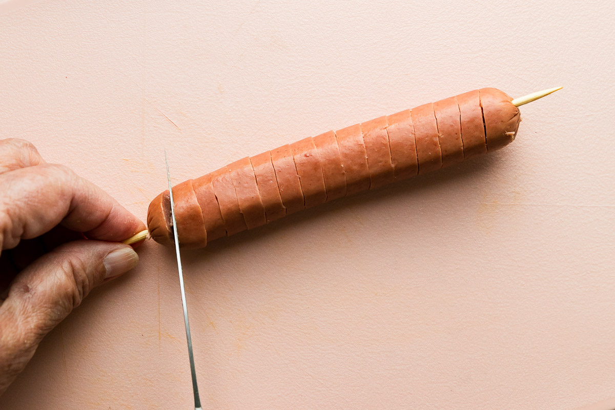A woman stabilizes a single hot dog with a wooden skewer placed through it lengthwise on a plastic pink cutting board with one hand while holding a pairing knife in the other. She presses the blade of the knife through the hot dog until it hits the skewer, while rotating the skewer, to create a spiral cut hot dog.