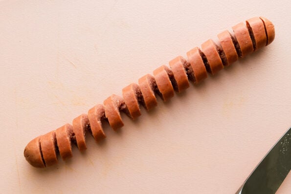 A spiral hot dog sits atop a pink plastic cutting board. A small pairing knife rests just below the hot dog.