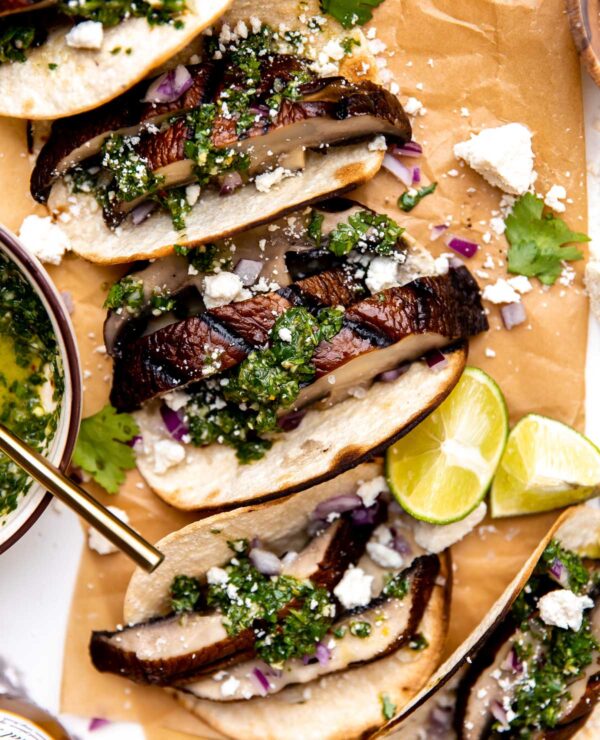 Assembled portobello mushroom tacos arranged on a piece of brown parchment paper. The parchment paper sits atop a white and gray marble surface. A ceramic bowl filled with chimichurri sauce, a small wooden pinch bowl filled with kosher salt, lime wedges, and a bottle of Modelo beer surrounds the grilled mushroom tacos at center.