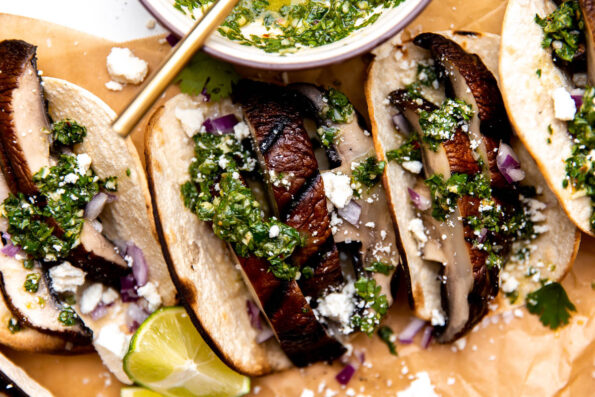 Assembled portobello mushroom tacos arranged on a piece of brown parchment paper. The parchment paper sits atop a white and gray marble surface. A ceramic bowl filled with chimichurri sauce rests alongside the grilled mushroom tacos at center with a gold spoon resting inside.