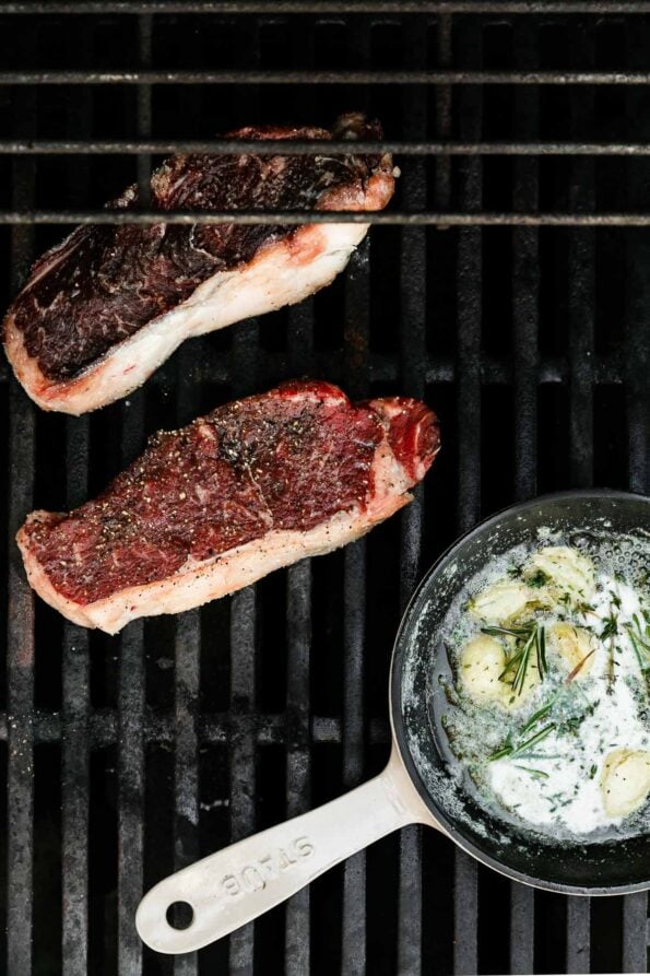 Two seasoned steaks sit atop a gas grill over indirect heat while a cream colored mini Staub cast iron fry pan is arranged alongside them melting garlic butter with a few herbs.