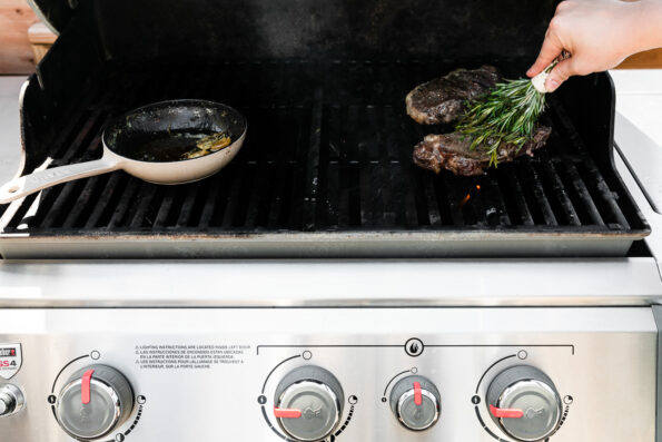 A woman's hand holds an assembled herb brush and uses it to rub garlic butter and the herb brush against the steaks grilling atop the gas grill grates. A cream colored mini Staub cast iron frying pan rests atop the grill grates on the other side of the grill.