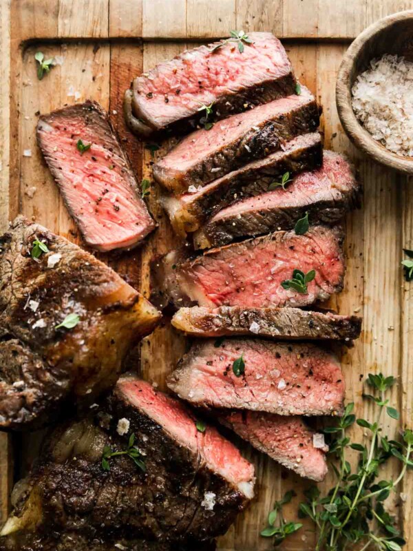How to grill steak, step 6: rest, carve, and serve. Grilled steak rests atop a wood cutting board and has been sliced to reveal medium rare doneness. The steak has been garnished with fresh herbs and a smoky finishing salt. A small wooden pinch bowl filled with smoky finishing salt and loose fresh herbs rest alongside the sliced strips of grilled steak and the cutting board sits atop a creamy white textured surface.