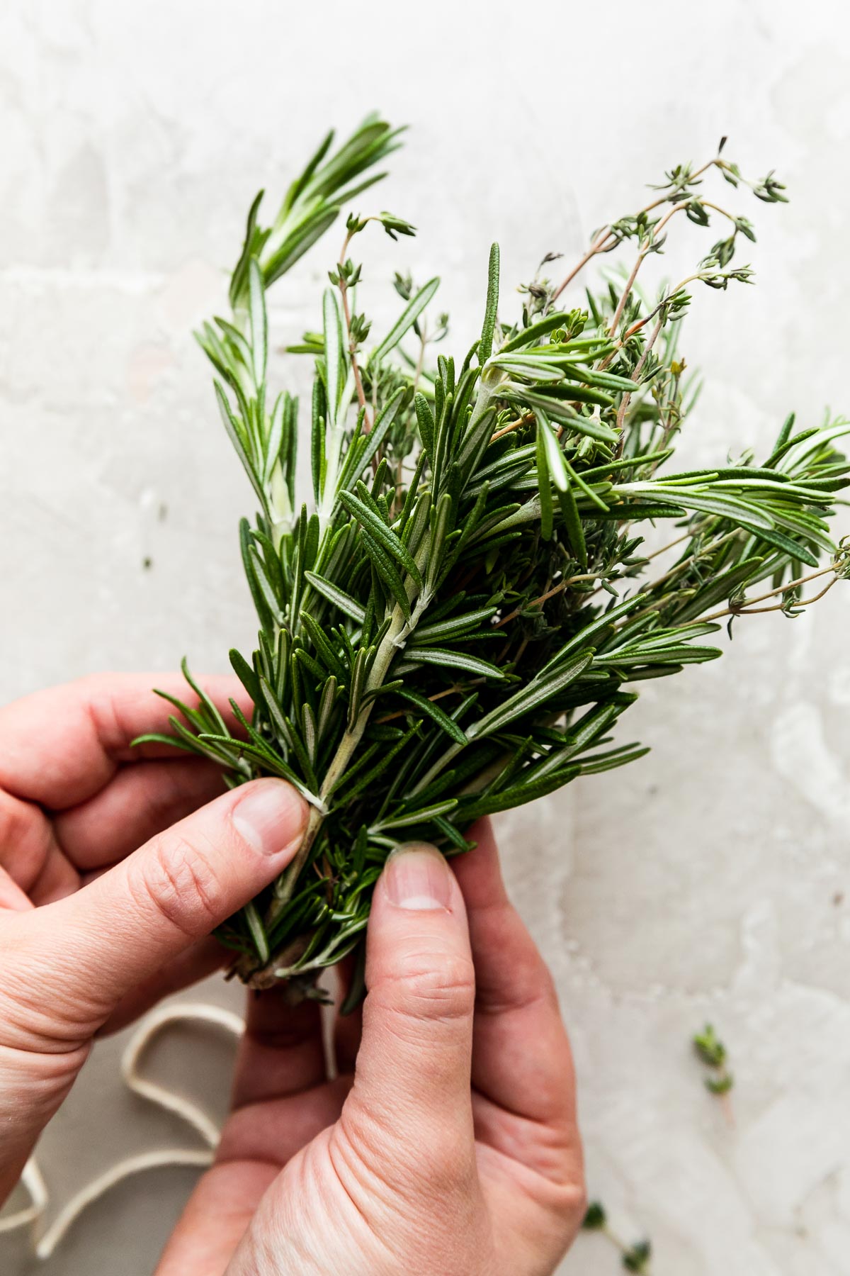 A woman's hands gather a large bunch of loose fresh rosemary and fresh thyme sprigs so that the ends of the sprigs are aligned. Her hands hold the gathered bunch of herbs above a creamy white textured surface. A single piece of kitchen twine rests on the surface below.