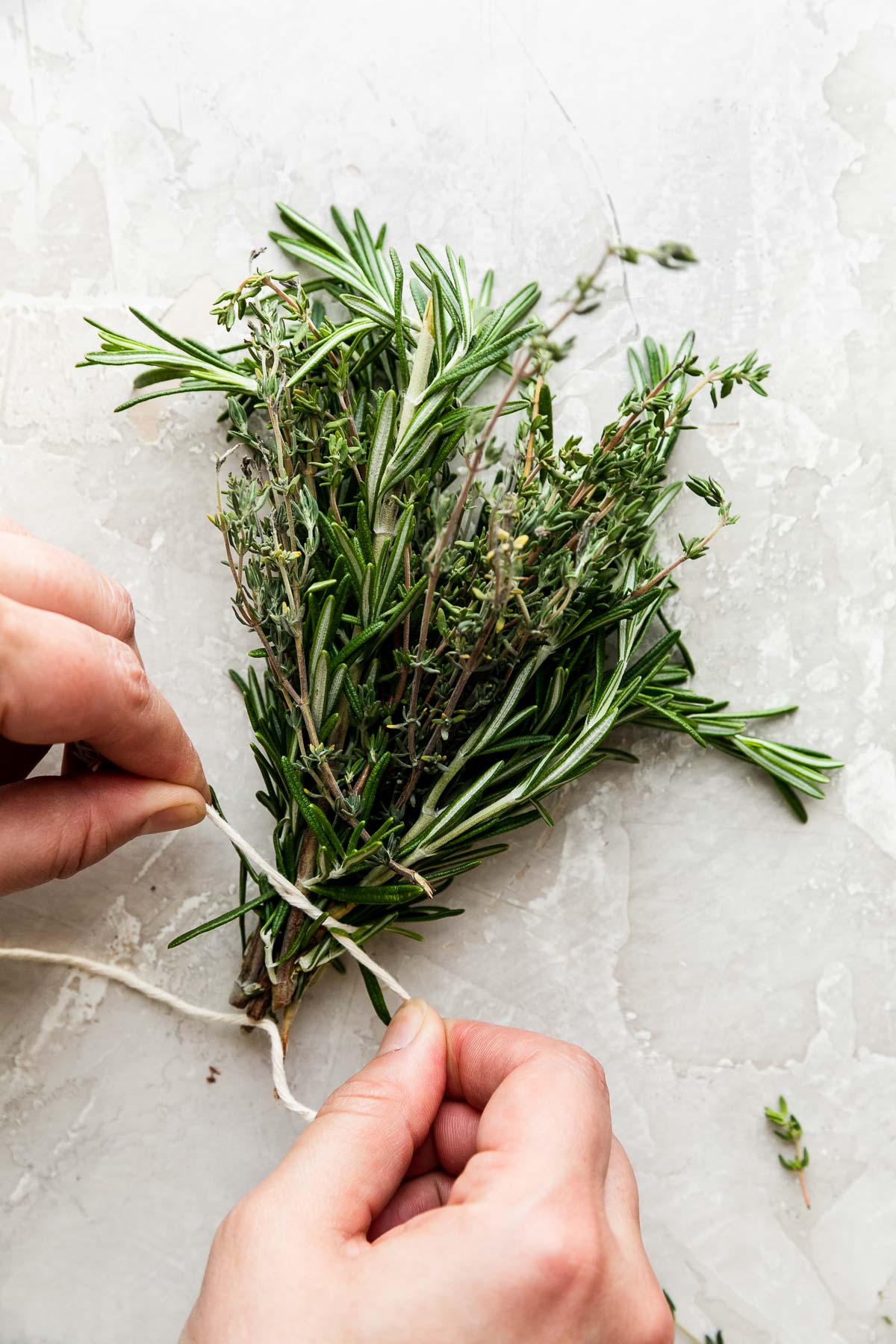 A bundle of gathered loose fresh rosemary and fresh thyme sprigs rest atop a creamy white textured surface. A woman's hands work to tie a piece of kitchen twine around the bottom of the herb bundle, securely.