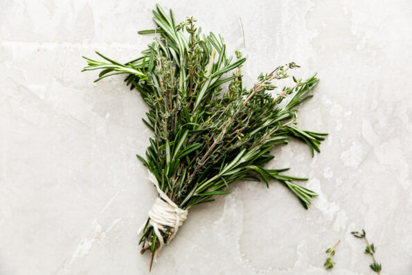 An assembled herb brush consisting of fresh rosemary and fresh thyme rests atop a creamy white textured surface. A few lose sprigs of thyme rest alongside the herb brush.