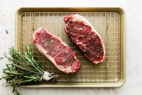 Two seasoned steaks arranged on a small gold baking sheet with a rack. An herb brush made with fresh herbs tied together with kitchen twine rests alongside the steaks and the baking sheet sits atop a creamy white textured surface and will be used while grilling steak.