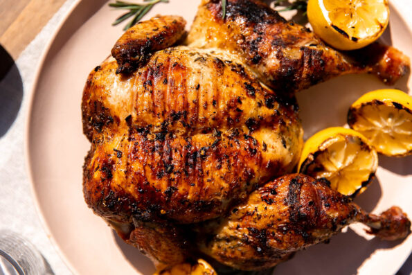 A finished tuscan grilled chicken rests atop a peach colored serving platter with fresh sprigs of rosemary and charred lemons surrounding the chicken. The platter sits atop a light gray linen table cloth.