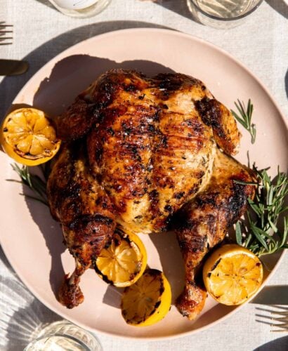A finished grilled spatchcock chicken rests atop a peach colored serving platter with fresh sprigs of rosemary and charred lemons surrounding the chicken. The platter sits atop a light gray and white linen table runner. Two glasses of wine, a white wine bottle, slices of fresh bread, a platter of grilled asparagus, and place settings surround the chicken at center.
