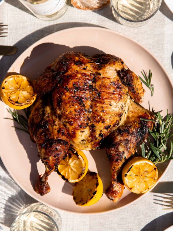 A finished grilled spatchcock chicken rests atop a peach colored serving platter with fresh sprigs of rosemary and charred lemons surrounding the chicken. The platter sits atop a light gray and white linen table runner. Two glasses of wine, a white wine bottle, slices of fresh bread, a platter of grilled asparagus, and place settings surround the chicken at center.