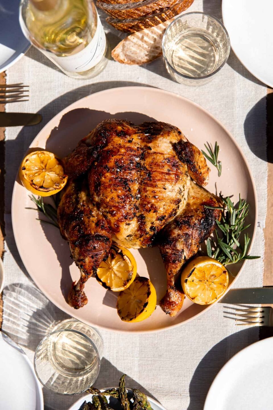 A finished grilled spatchcock chicken rests atop a peach colored serving platter with fresh sprigs of rosemary and charred lemons surrounding the chicken. The platter sits atop a light gray and white linen table runner. Two glasses of wine, a white wine bottle, slices of fresh bread, a platter of grilled asparagus, and place settings surround the chicken at center.