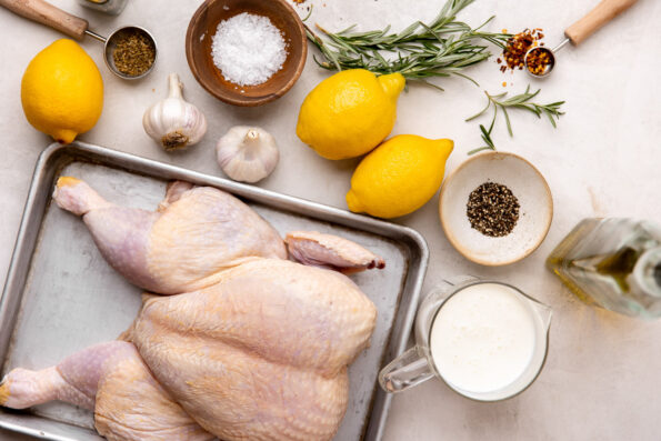 Tuscan grilled chicken ingredients arranged on a creamy white textured surface: whole chicken, olive oil, lemons, garlic, fresh rosemary, dried oregano, crushed red pepper flakes, buttermilk, kosher salt, and ground black pepper.