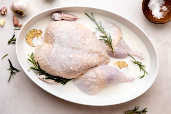 A whole chicken is submerged in a shallow serving dish filled with buttermilk, salt, garlic, and rosemary for brining. The dish rests atop a creamy white textured surface with two whole bulbs of garlic, a few loose garlic cloves, a small wooden pinch bowl filled with flaky salt, and a few loose sprigs of fresh rosemary surrounding.