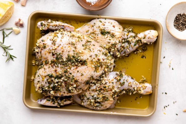 How to make grilled spatchcock chicken, step 2: marinate the chicken. A whole chicken rests atop a small gold baking dish. The chicken has been spatchcocked to lay flat & is arranged breast-side up on the baking sheet. A Tuscan marinade has been rubbed all over the chicken to be prepped for grilling. The baking sheet sits atop a creamy white textured surface with a bulb of garlic with loose garlic cloves, a discarded half of a zested & juice lemon, a few loose sprigs of fresh rosemary, a small wooden pinch bowl filled with flaky salt, and a small ceramic pinch bowl filled with ground black pepper surround the marinated chicken at center.