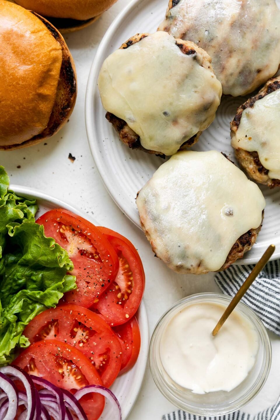 Four chicken burgers patties topped with melted cheese sit atop a white plate. Another white ceramic plate filled with lettuce, sliced tomatoes, and sliced onion sits adjacent to the plate of grilled burgers. The two plates are surrounded by toasted buns, a small clear glass jar filled with mayonnaise and a small gold spoon. A blue and white striped linen napkin rests alongside the plate.
