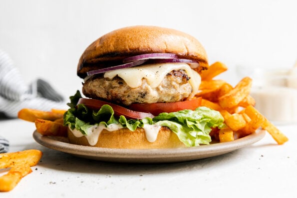 Grilled chicken burger is served on a small ceramic plate with french fries. The plate sits atop a creamy white textured surface with a blue and white striped linen napkin tucked alongside. A small glass jar filled with mayonnaise sits in the background out of focus.