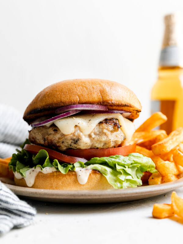Grilled chicken burger is served on a small ceramic plate with french fries. The plate sits atop a creamy white textured surface with a blue and white striped linen napkin tucked alongside. A bottle of beer sits in the background out of focus.