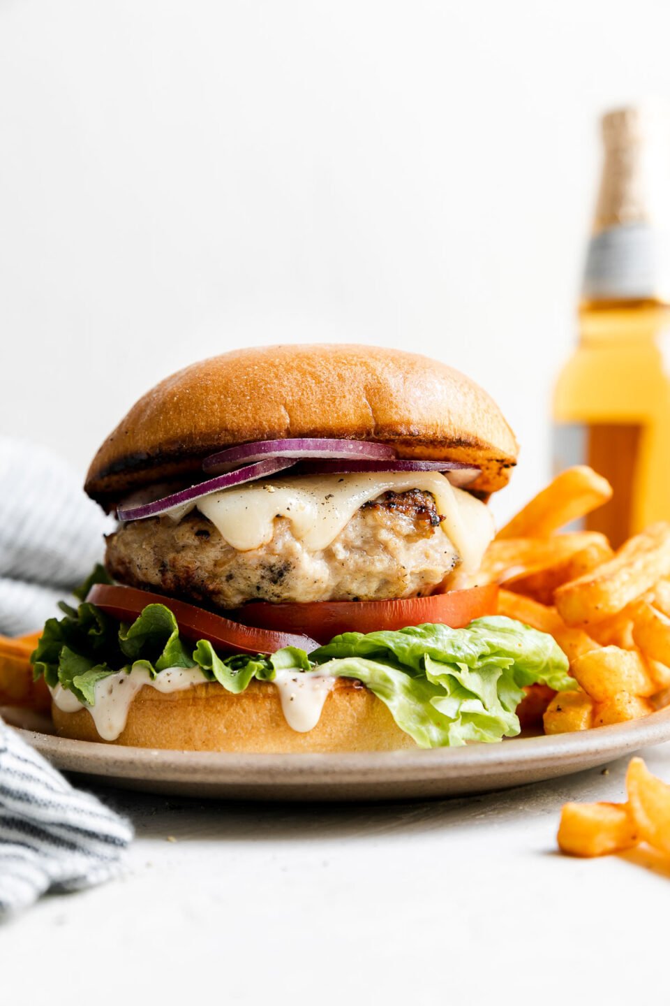 Grilled chicken burger is served on a small ceramic plate with french fries. The plate sits atop a creamy white textured surface with a blue and white striped linen napkin tucked alongside. A bottle of beer sits in the background out of focus.