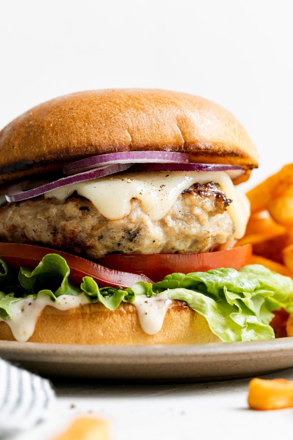 A single grilled chicken burger is served on a small ceramic plate with french fries atop a creamy white textured surface with a blue and white striped linen napkin tucked alongside.