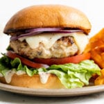 A single grilled chicken burger is served on a small ceramic plate with french fries. The plate sits atop a creamy white with a small glass jar filled with mayonnaise and a small gold spoon resting inside.