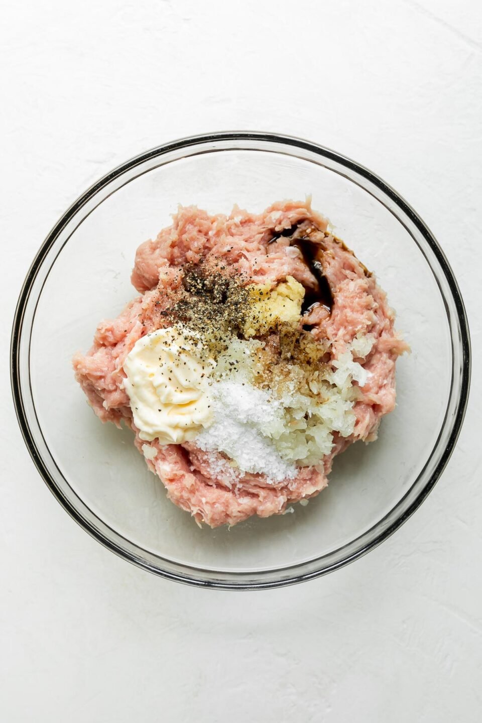 Ground chicken, yellow onion, garlic, mayonnaise, and Worcestershire sauce inside of a clear glass mixing bowl atop a creamy white textured surface.