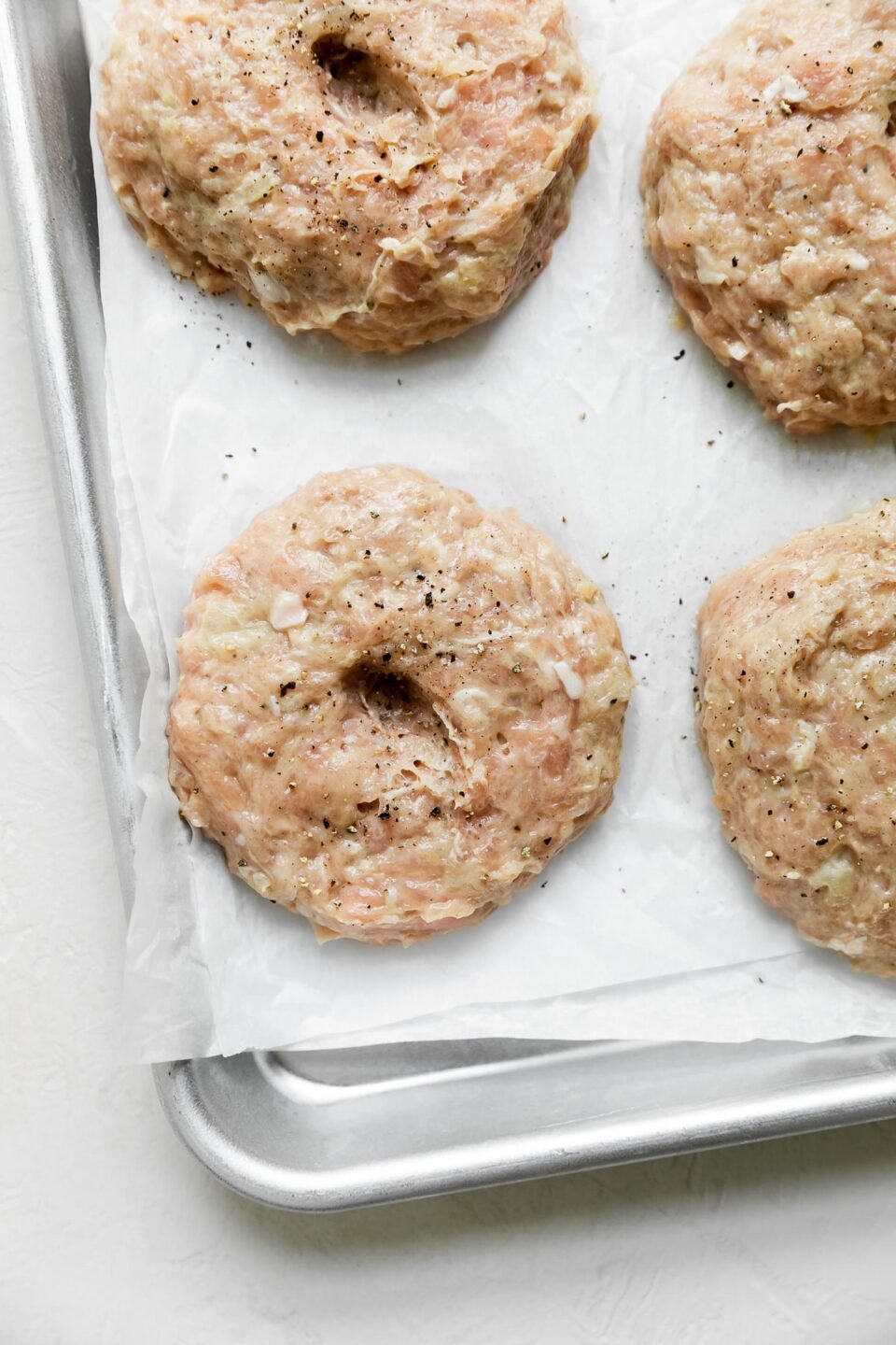 Four raw and uncooked chicken burger patties placed on a small aluminum baking sheet atop crumpled parchment paper. The baking sheet sits atop a creamy white textured surface.
