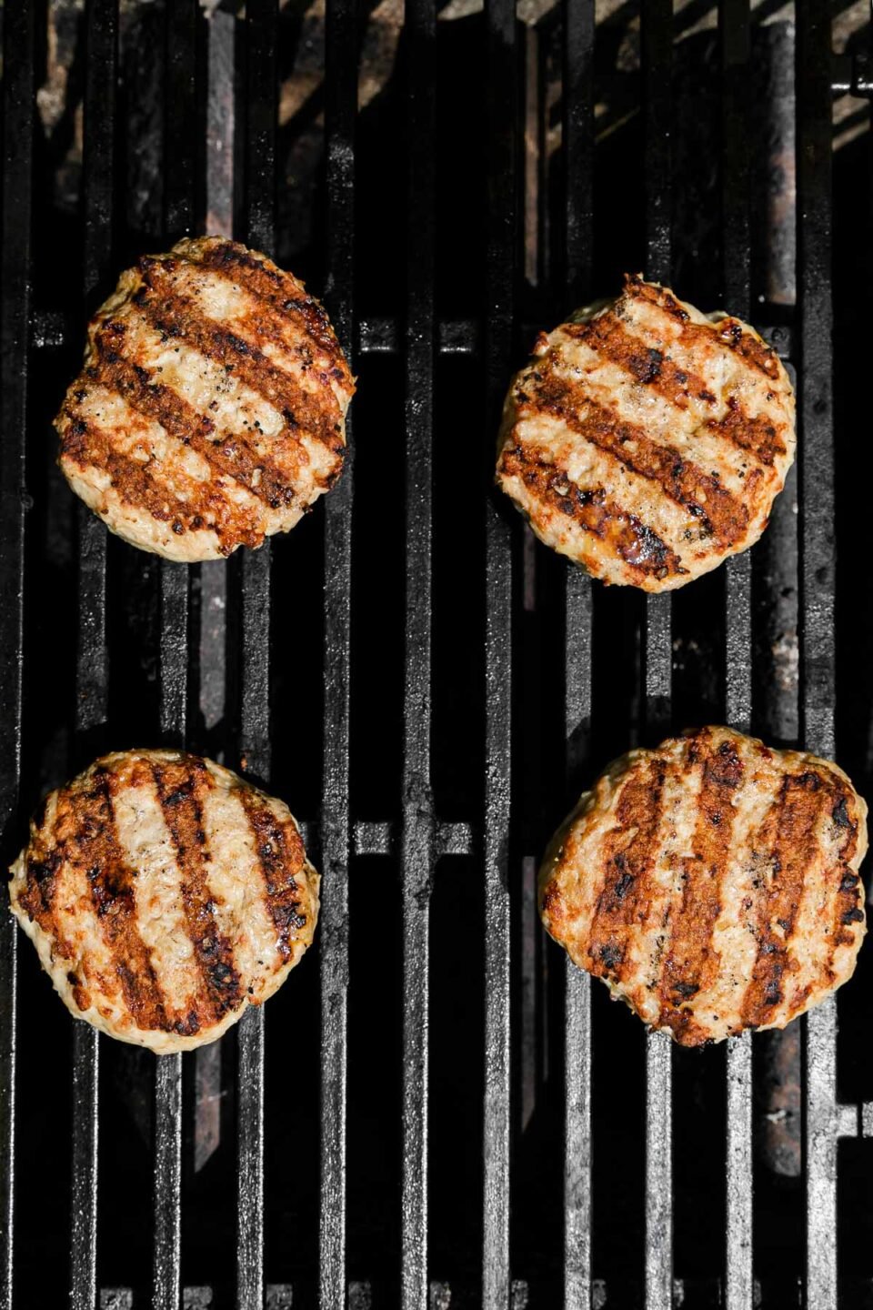 Four chicken burger patties with grill marks sit atop gas grill grates while being cooked.